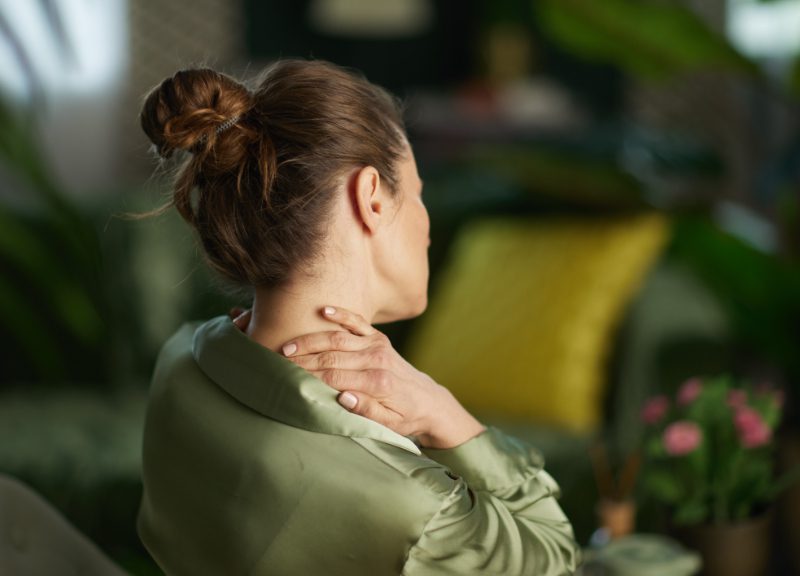 A woman in a green blouse reaches back to touch her neck, depicting muscle tension and the need for a mindful pause and physical relief in her home environment.